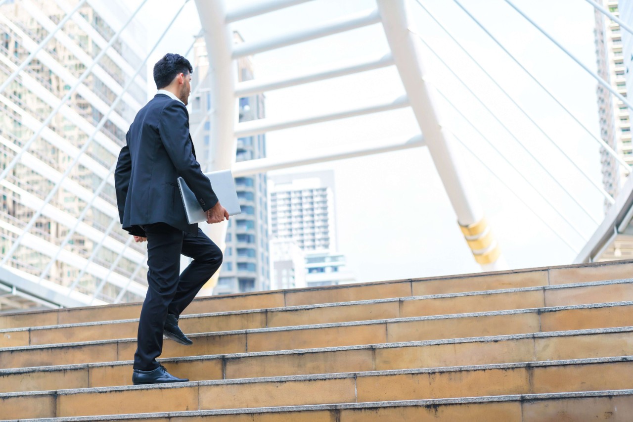 Man walking up the stairs holding a laptop computer while standing outdoors with office buildings in the background Man walking up the stairs holding a laptop computer while standing outdoors with office buildings in the background