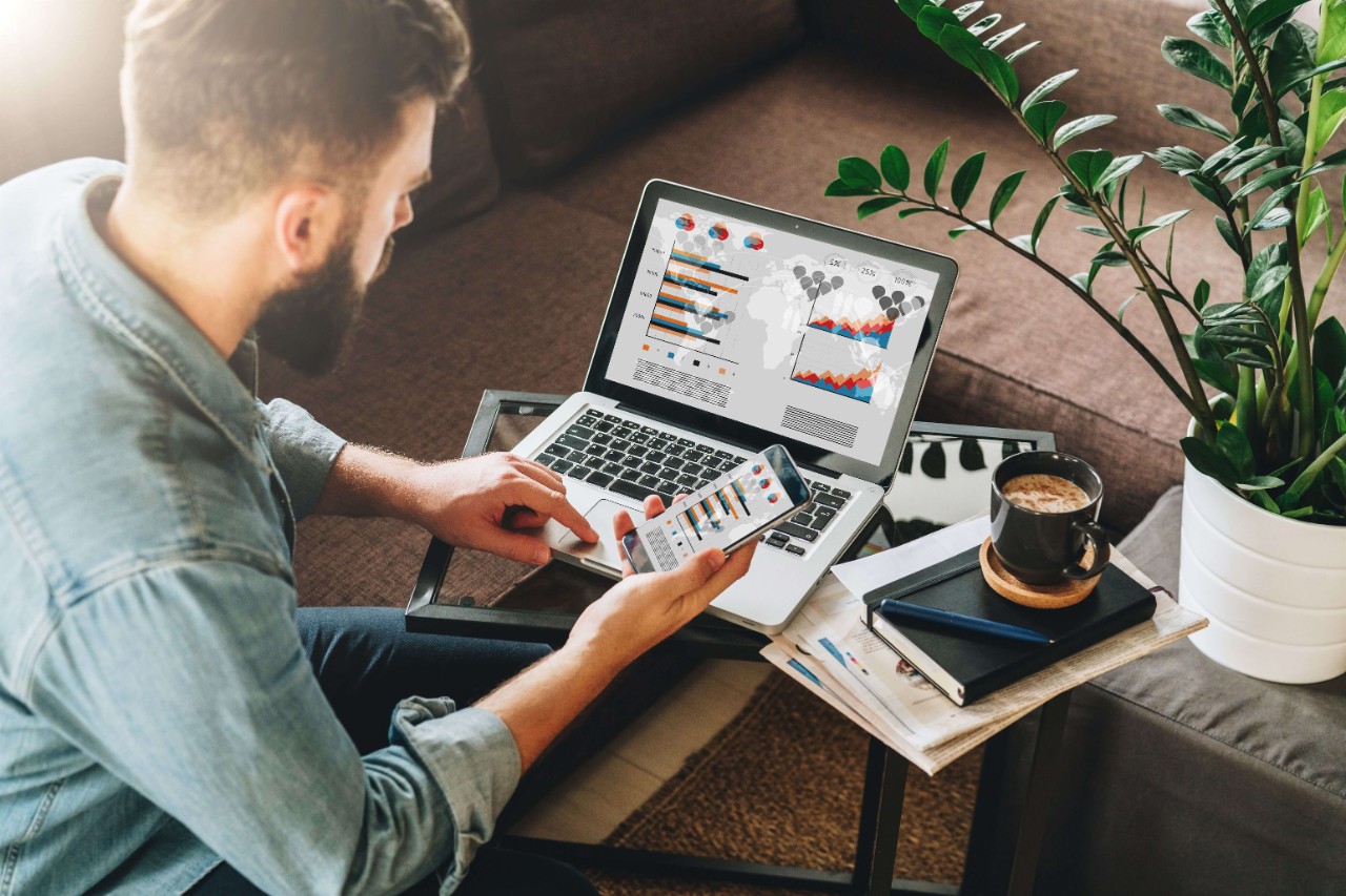 Man, entrepreneur, freelancer sits at home on couch at coffee table, uses smartphone, working on laptop with graphs Man, entrepreneur, freelancer sits at home on couch at coffee table, uses smartphone, working on laptop with graphs